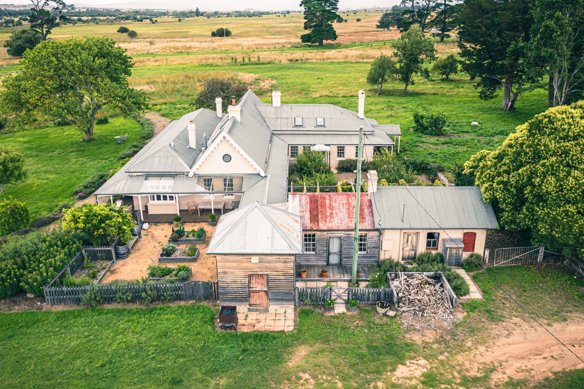 Aerial view of the Mill Pond Farm homestead at Jembaicumbene, showing hipped grey roofs, chimneys, a corrugated iron outbuilding, a timber barn, a vegetable garden, and surrounding green paddocks.