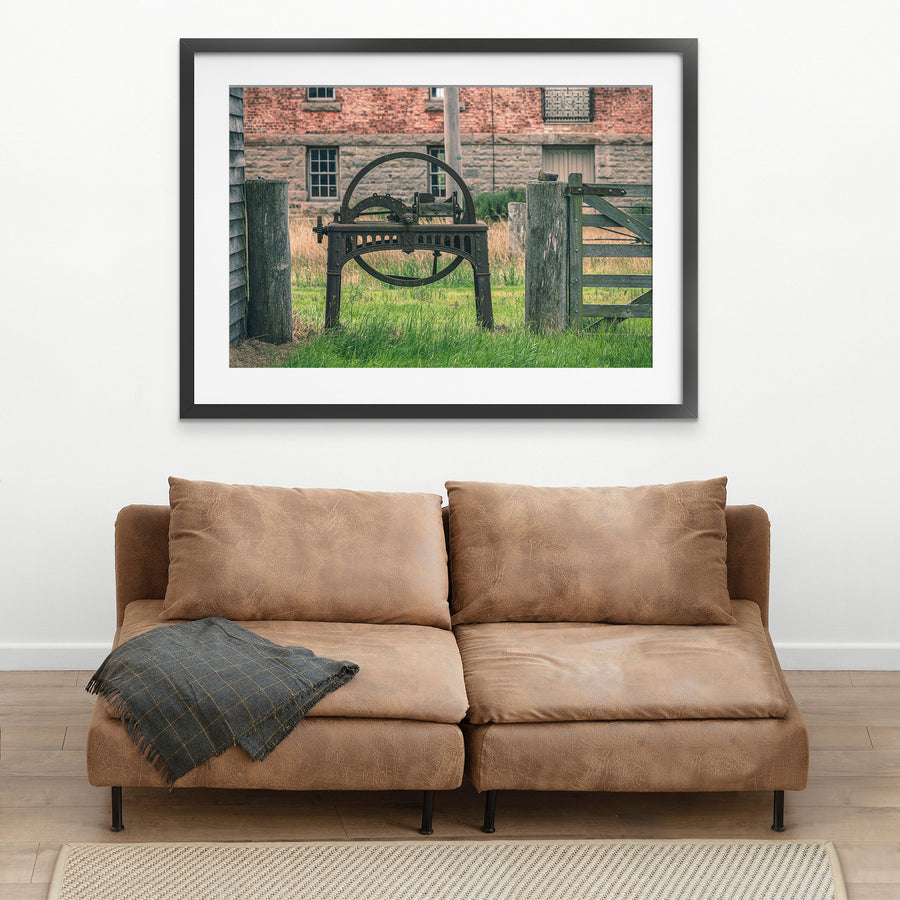 A rusting cast iron flywheel cutter with a large spoked wheel and curved toothed arc stands in long grass beside a wooden farm gate, with the brick and stone mill building at Jembaicumbene visible behind.