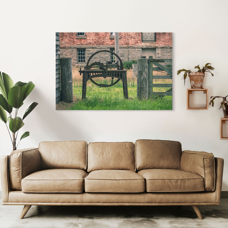 A rusting cast iron flywheel cutter with a large spoked wheel and curved toothed arc stands in long grass beside a wooden farm gate, with the brick and stone mill building at Jembaicumbene visible behind.