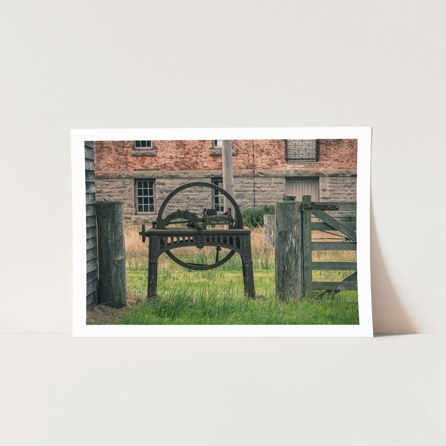 A rusting cast iron flywheel cutter with a large spoked wheel and curved toothed arc stands in long grass beside a wooden farm gate, with the brick and stone mill building at Jembaicumbene visible behind.