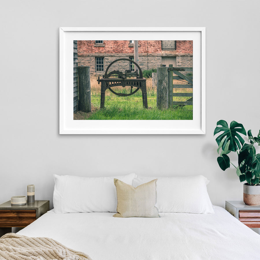 A rusting cast iron flywheel cutter with a large spoked wheel and curved toothed arc stands in long grass beside a wooden farm gate, with the brick and stone mill building at Jembaicumbene visible behind.