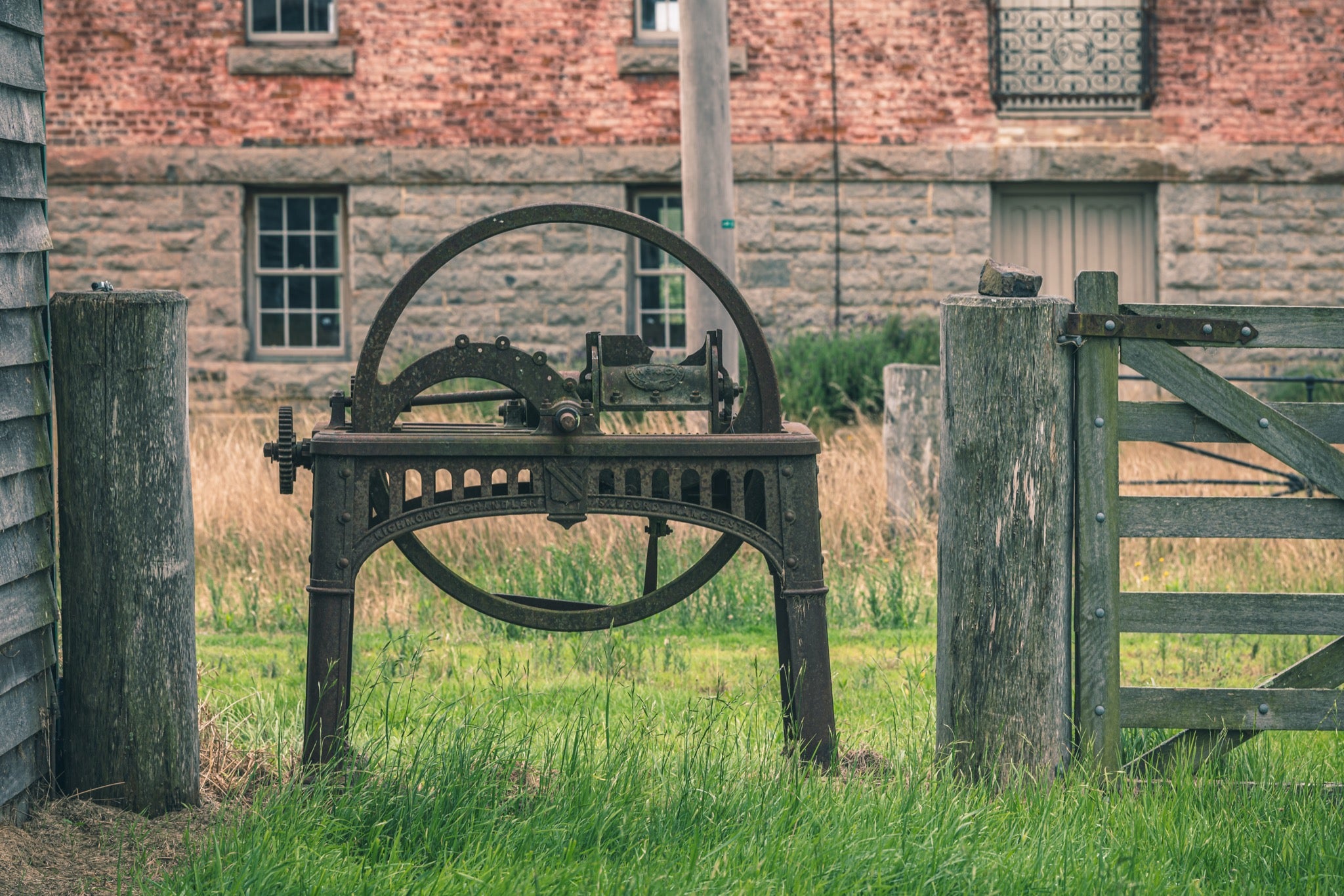 A rusting cast iron flywheel cutter with a large spoked wheel and curved toothed arc stands in long grass beside a wooden farm gate, with the brick and stone mill building at Jembaicumbene visible behind.