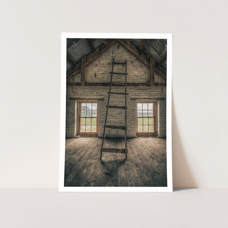 A timber ladder leans against a whitewashed brick gable wall rising to the roof apex inside the mill at Jembaicumbene, with two red-framed sash windows open to fields beyond.
