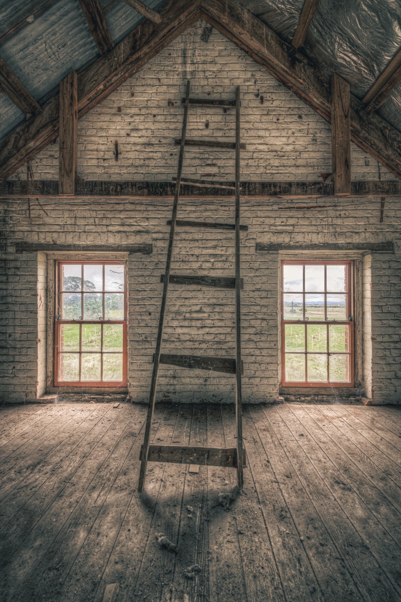 A timber ladder leans against a whitewashed brick gable wall rising to the roof apex inside the mill at Jembaicumbene, with two red-framed sash windows open to fields beyond.