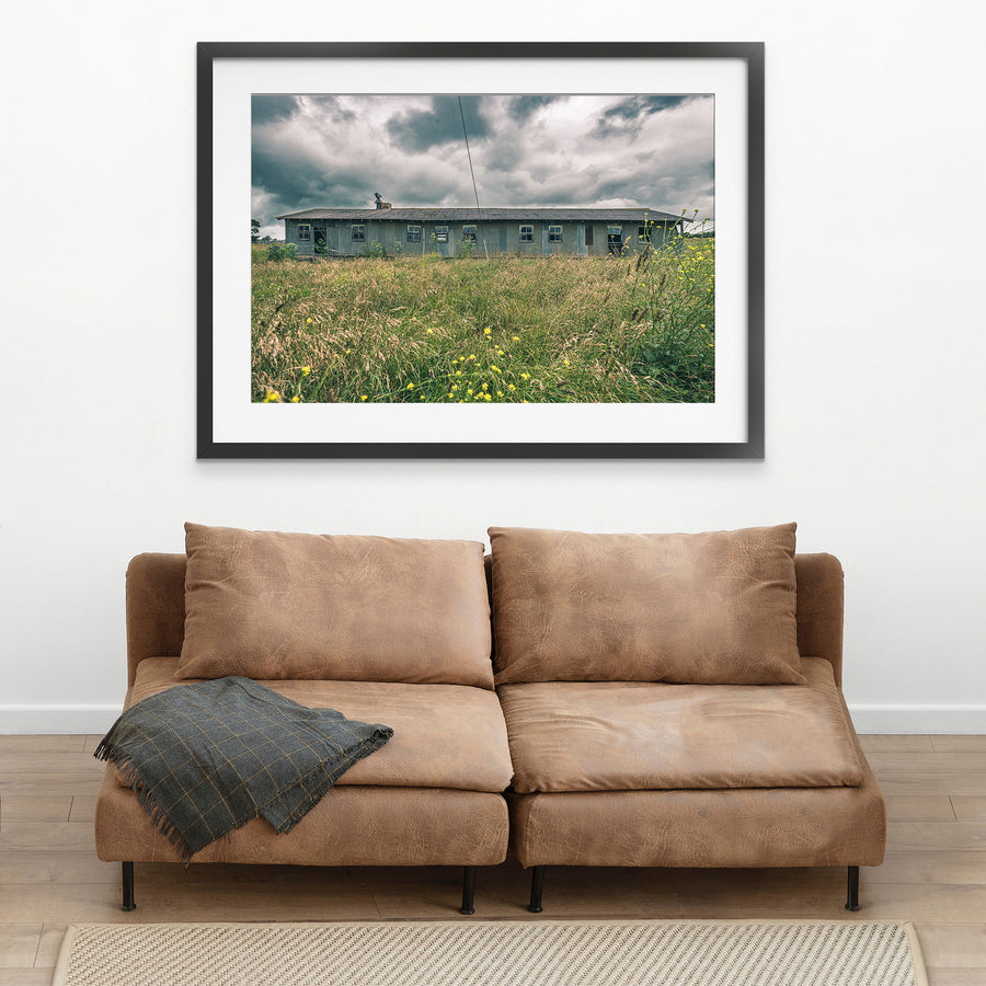 A long, weathered corrugated iron shed with timber-framed windows stands behind tall summer grass and yellow wildflowers at Jembaicumbene, New South Wales, under an overcast sky.