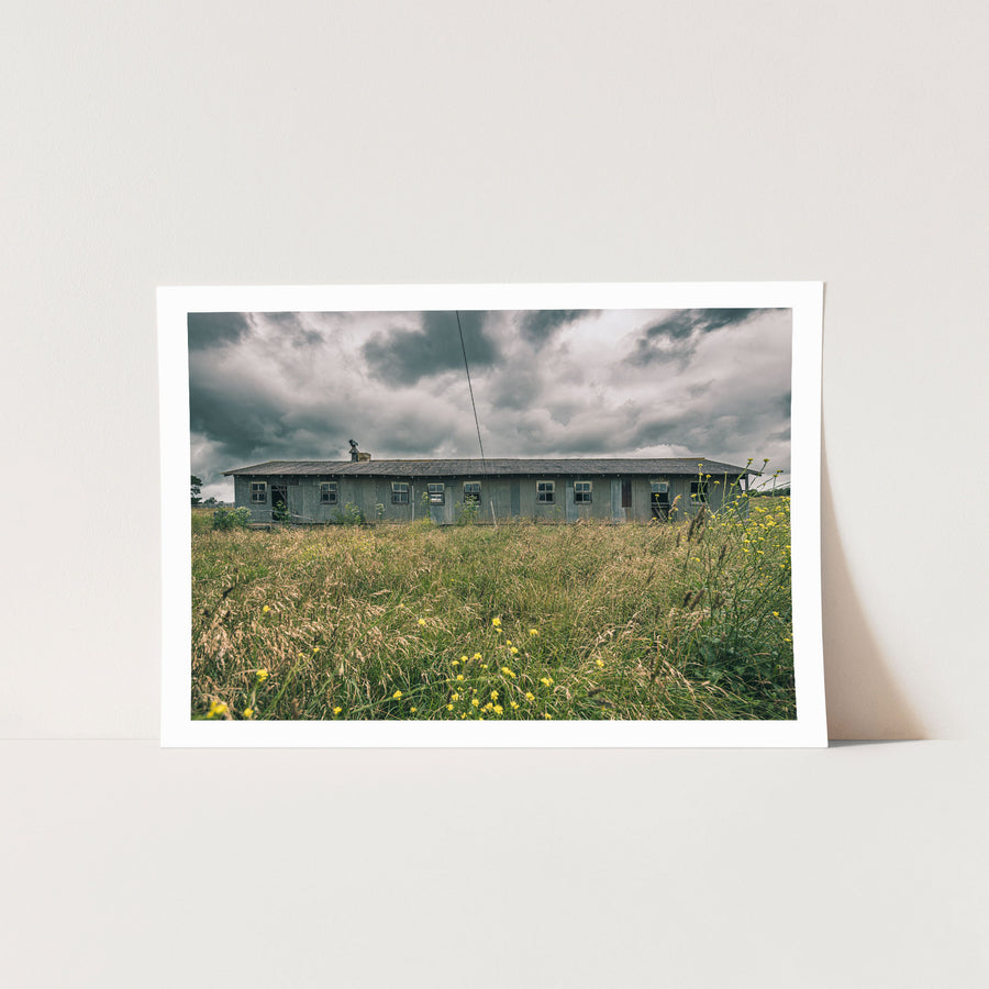 A long, weathered corrugated iron shed with timber-framed windows stands behind tall summer grass and yellow wildflowers at Jembaicumbene, New South Wales, under an overcast sky.