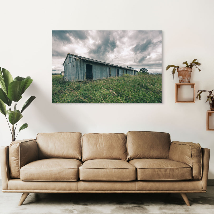 A weathered corrugated iron building with empty window openings and an open doorway stands in tall grass at Jembaicumbene, New South Wales.