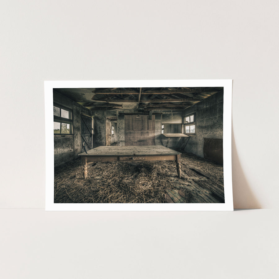 A long timber table with turned legs stands on a straw-strewn floor inside a derelict outbuilding at Mill Pond Farm, Jembaicumbene, with exposed rafters overhead and light entering from windows on either side.