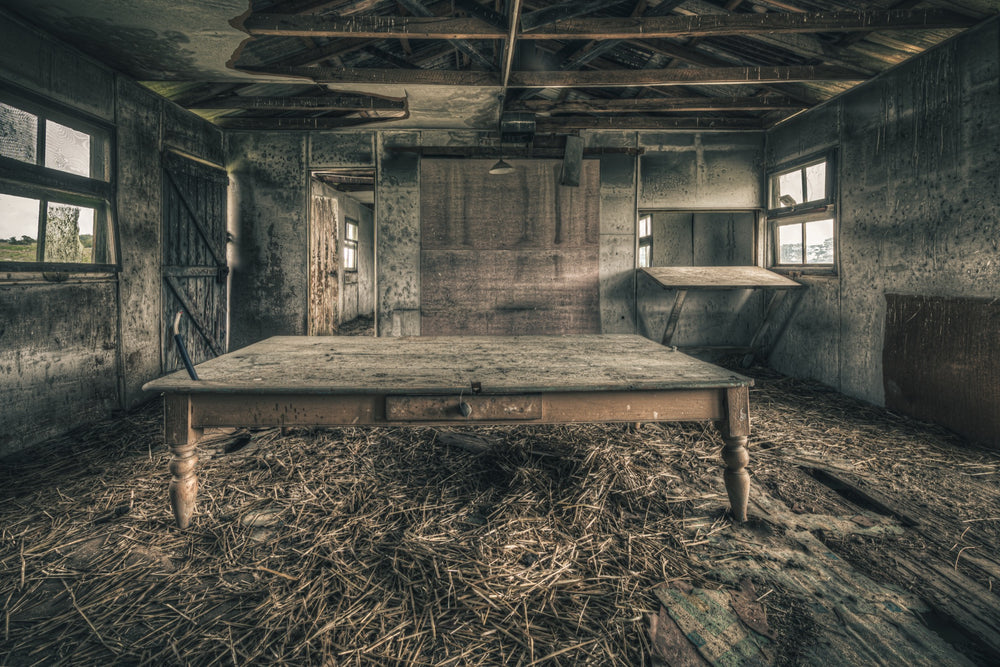 A long timber table with turned legs stands on a straw-strewn floor inside a derelict outbuilding at Mill Pond Farm, Jembaicumbene, with exposed rafters overhead and light entering from windows on either side.