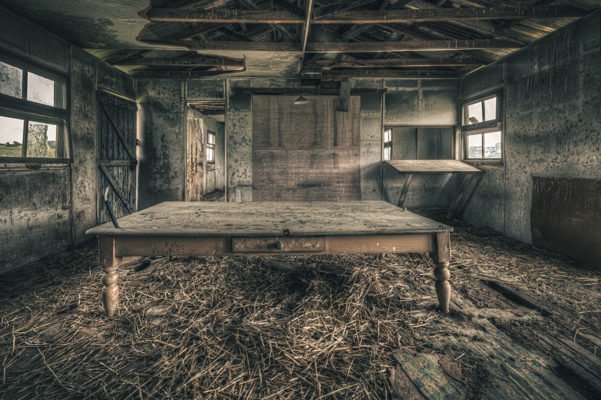 A long timber table with turned legs stands on a straw-strewn floor inside a derelict outbuilding at Mill Pond Farm, Jembaicumbene, with exposed rafters overhead and light entering from windows on either side.