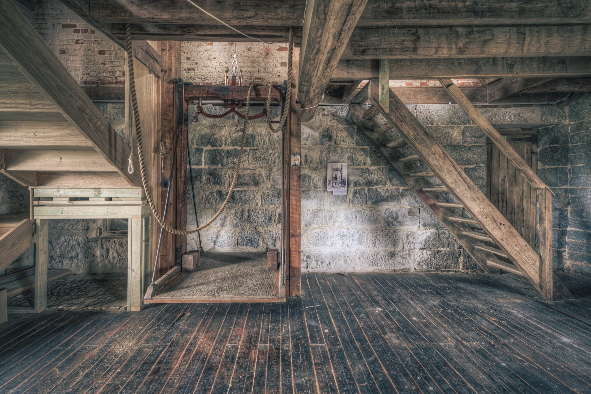 Timber staircase and rope hoist mechanism on the mill hoist floor at Jembaicumbene, with a stone block wall and heavy hardwood beams overhead.