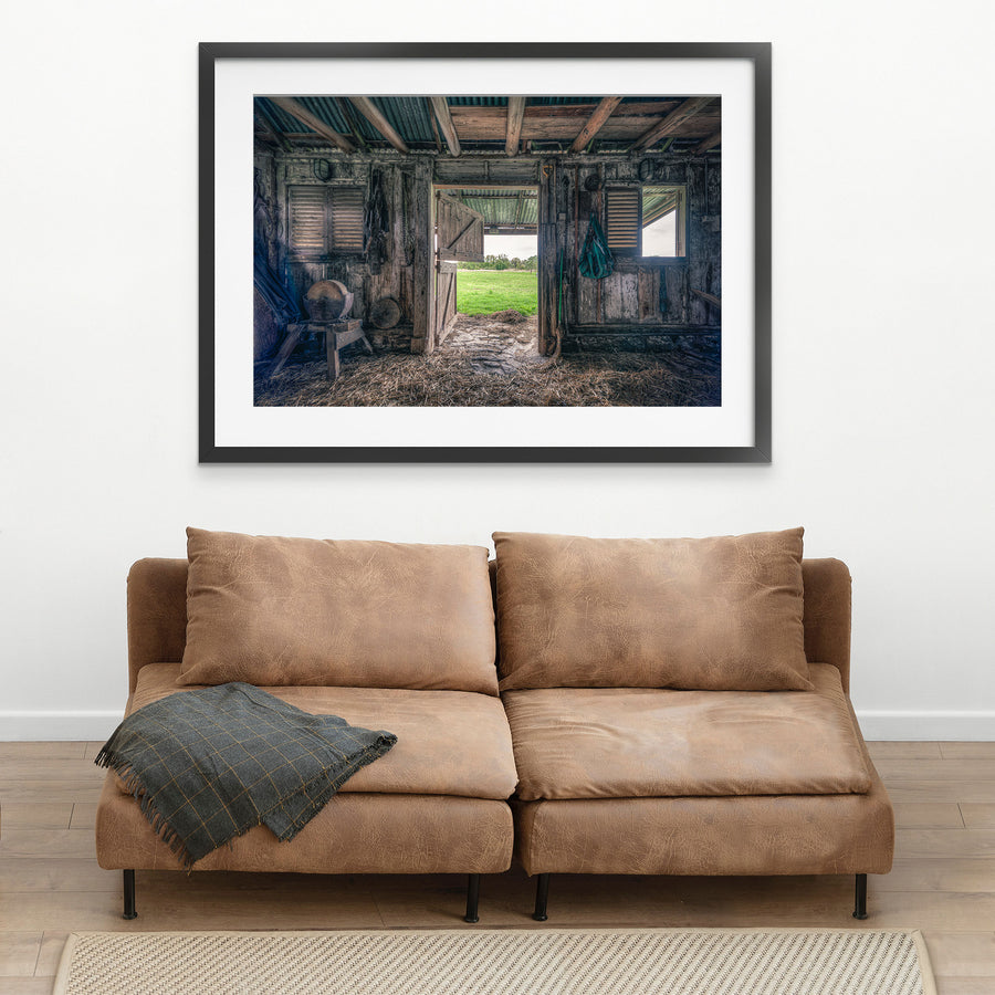 Interior of the 1840s timber wagon barn at Jembaicumbene, looking through an open doorway to a green paddock and treeline, with a chain and hay net hanging from weathered posts and straw on the dirt floor.