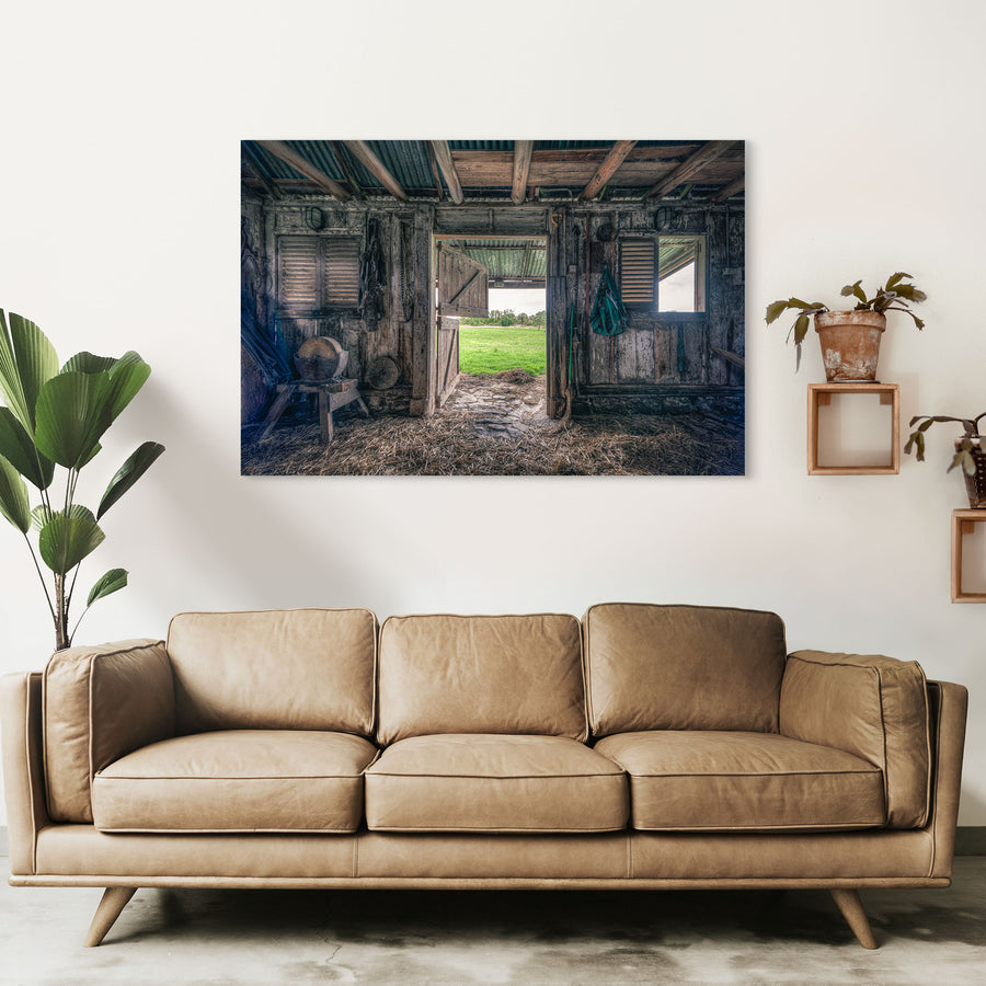 Interior of the 1840s timber wagon barn at Jembaicumbene, looking through an open doorway to a green paddock and treeline, with a chain and hay net hanging from weathered posts and straw on the dirt floor.