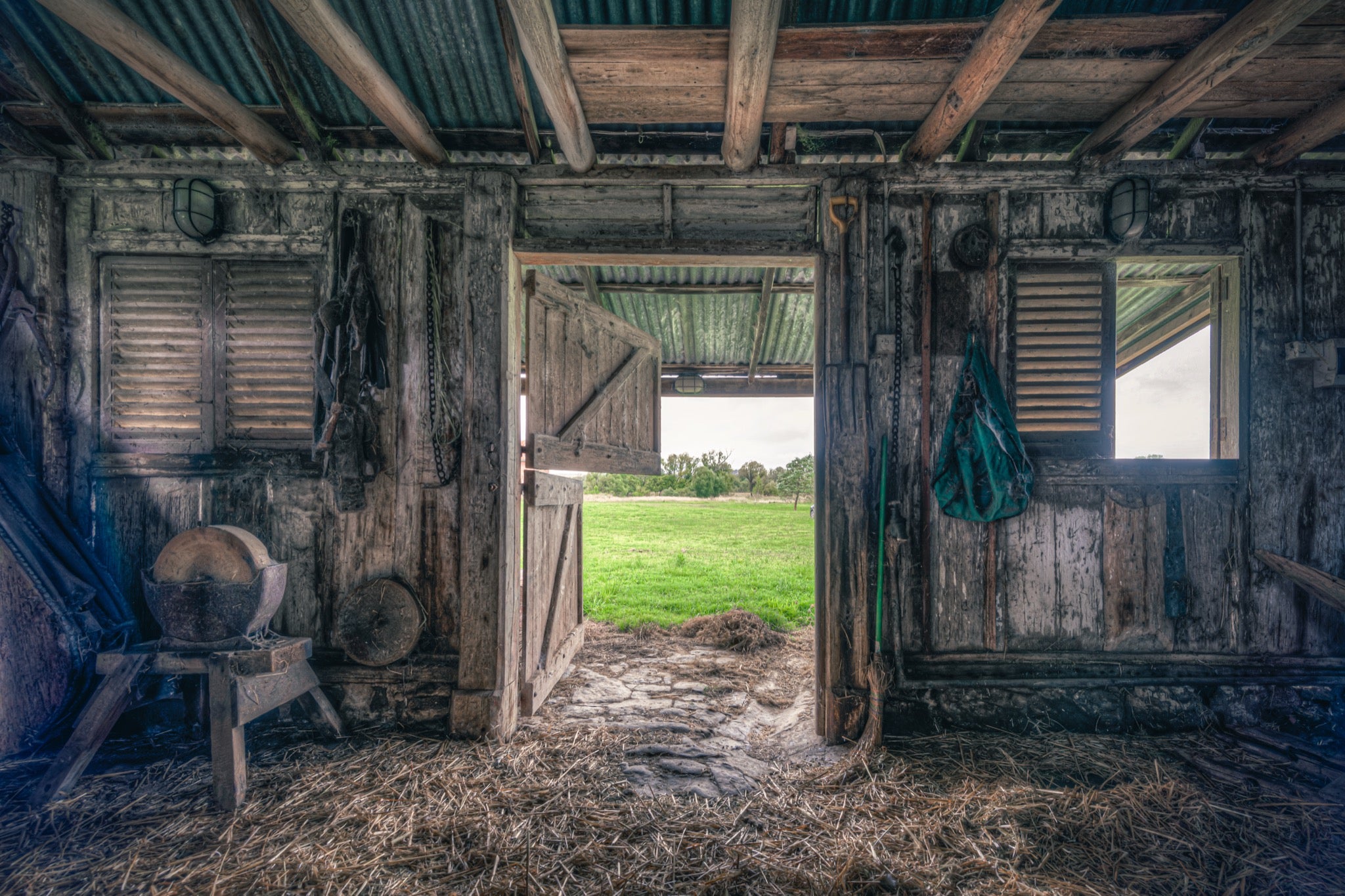 Interior of the 1840s timber wagon barn at Jembaicumbene, looking through an open doorway to a green paddock and treeline, with a chain and hay net hanging from weathered posts and straw on the dirt floor.
