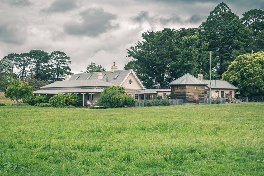 A weatherboard homestead with Gothic Revival bargeboards and a corrugated iron roof, a timber outbuilding to the right, and a broad green paddock in the foreground at Jembaicumbene, NSW.