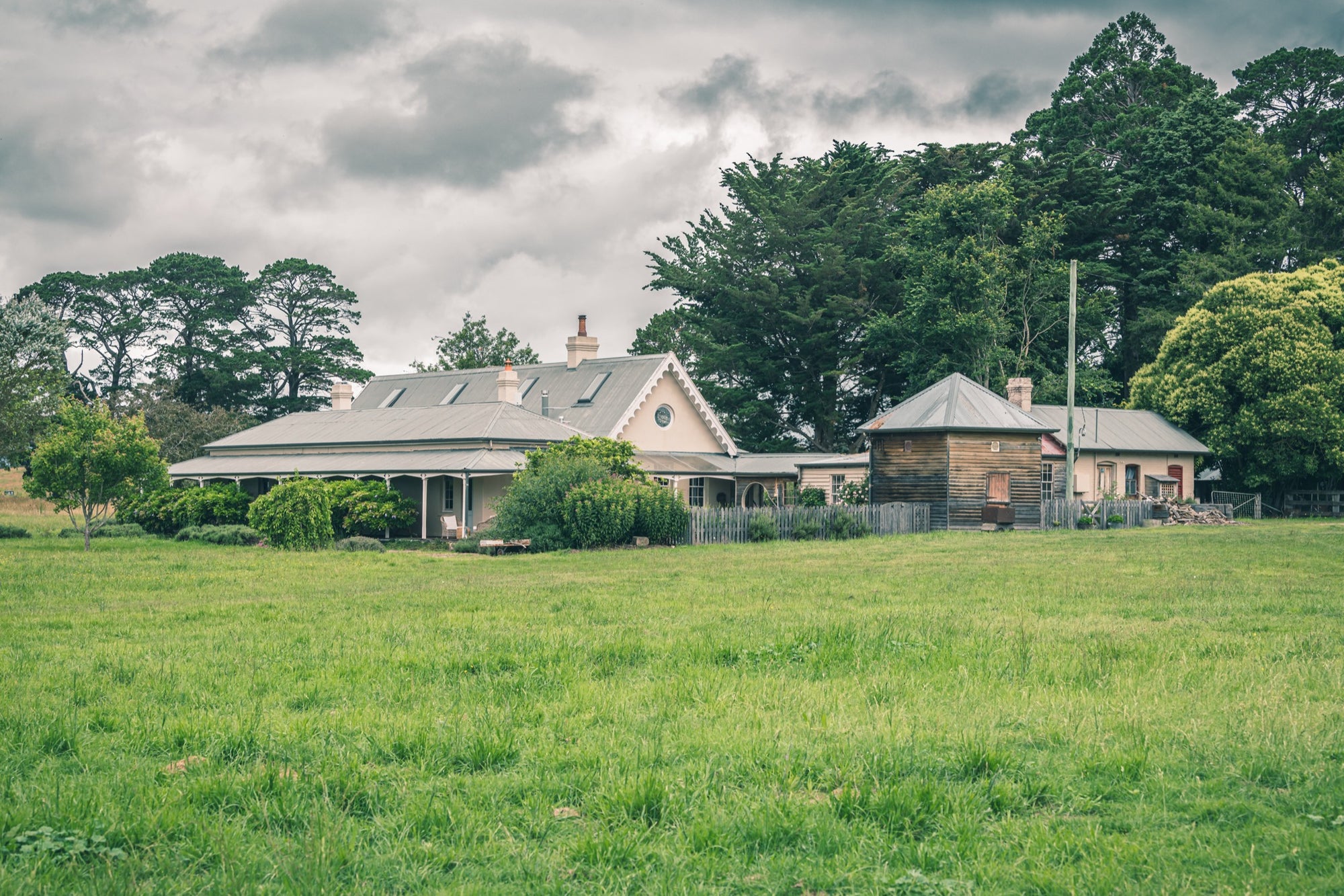 A weatherboard homestead with Gothic Revival bargeboards and a corrugated iron roof, a timber outbuilding to the right, and a broad green paddock in the foreground at Jembaicumbene, NSW.