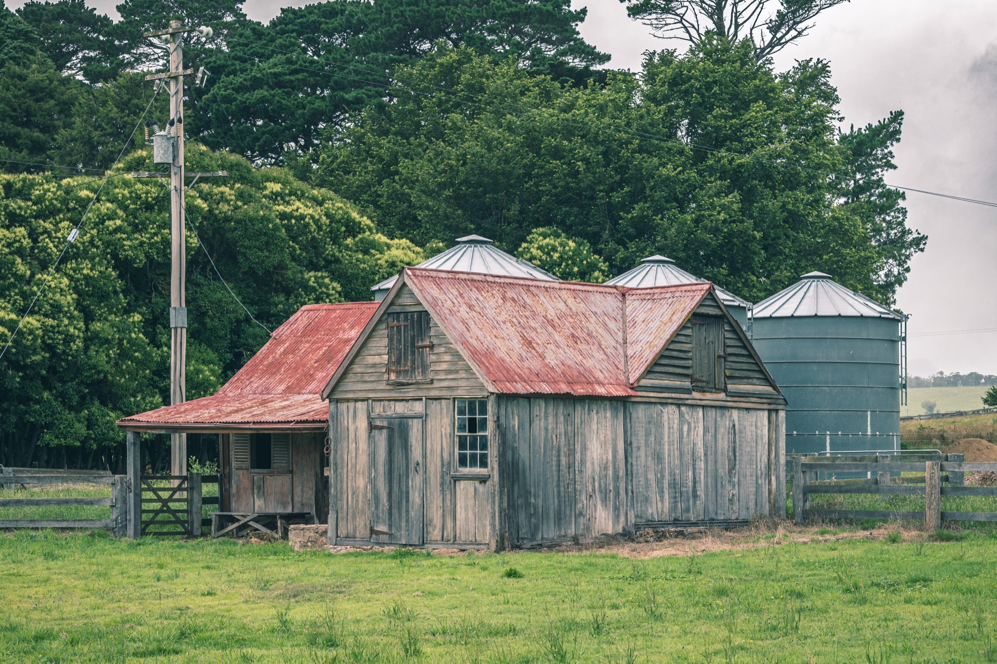 Two rusted corrugated iron-roofed timber sheds and galvanised grain silos behind a post-and-rail fence at Jembaicumbene, NSW.