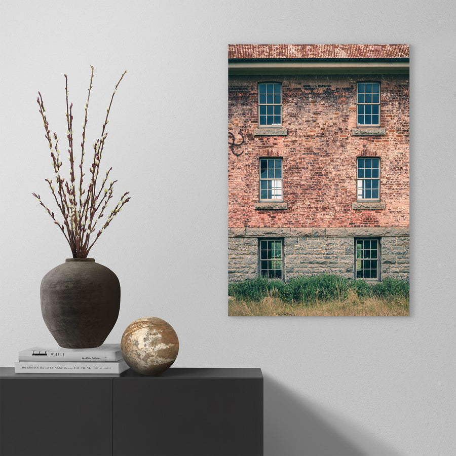 Six multi-pane sash windows set in three pairs across a brick and stone facade at Jembaicumbene, with corrugated metal roofing above and tall grass and purple flowering plants at the base.