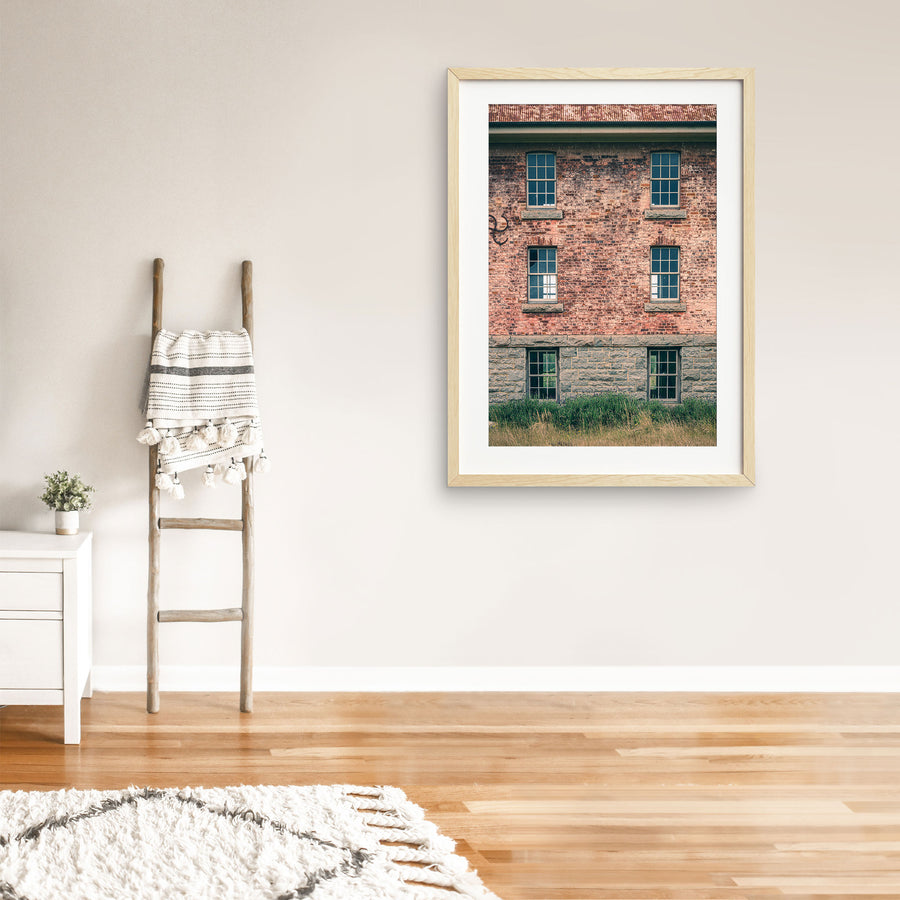 Six multi-pane sash windows set in three pairs across a brick and stone facade at Jembaicumbene, with corrugated metal roofing above and tall grass and purple flowering plants at the base.