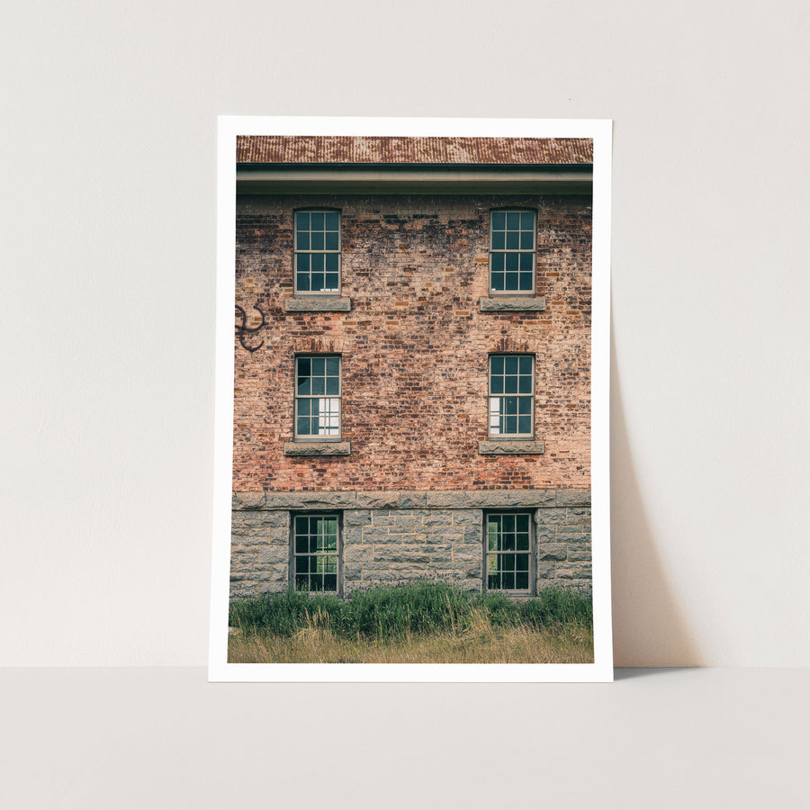Six multi-pane sash windows set in three pairs across a brick and stone facade at Jembaicumbene, with corrugated metal roofing above and tall grass and purple flowering plants at the base.