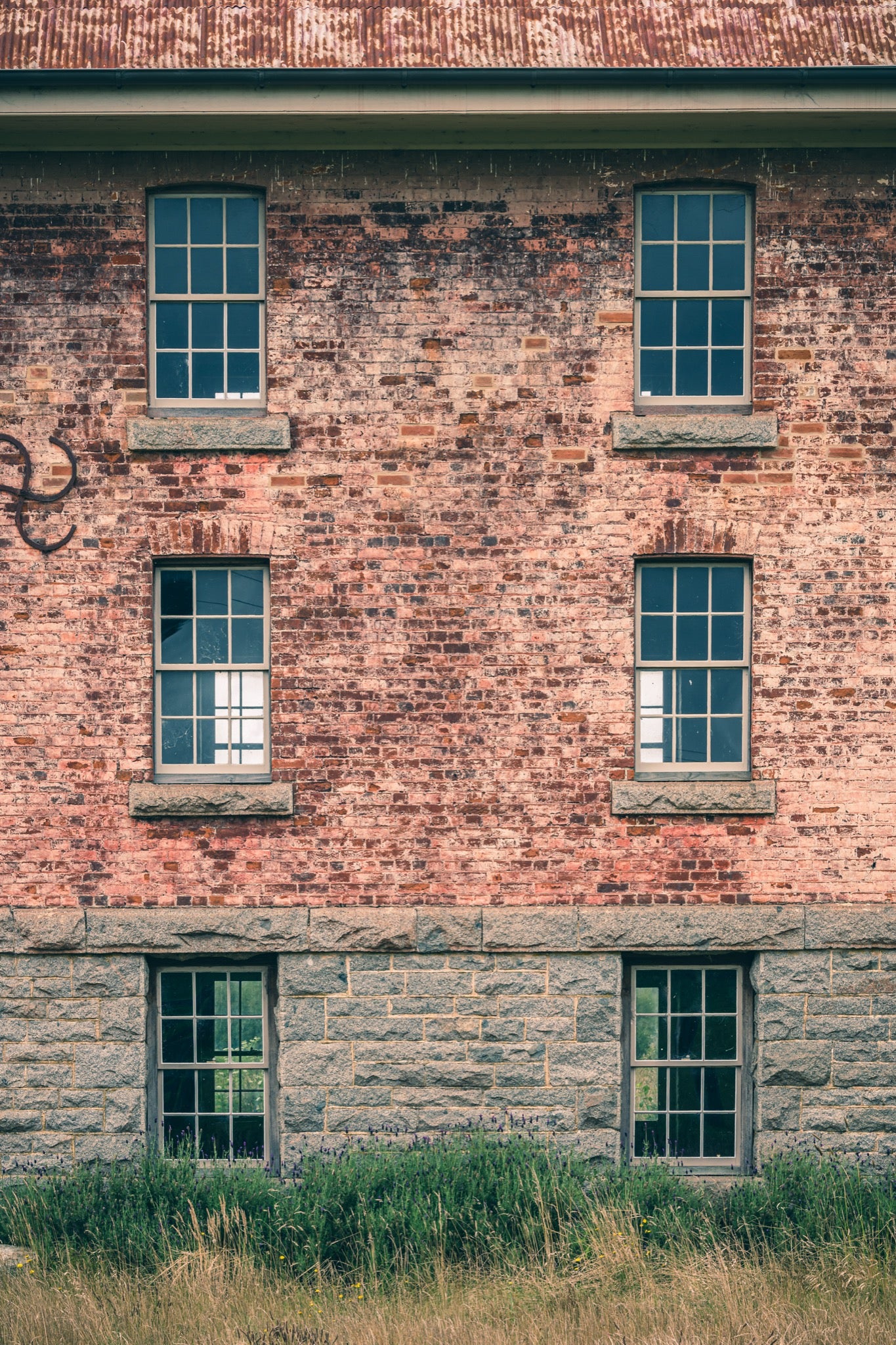 Six multi-pane sash windows set in three pairs across a brick and stone facade at Jembaicumbene, with corrugated metal roofing above and tall grass and purple flowering plants at the base.