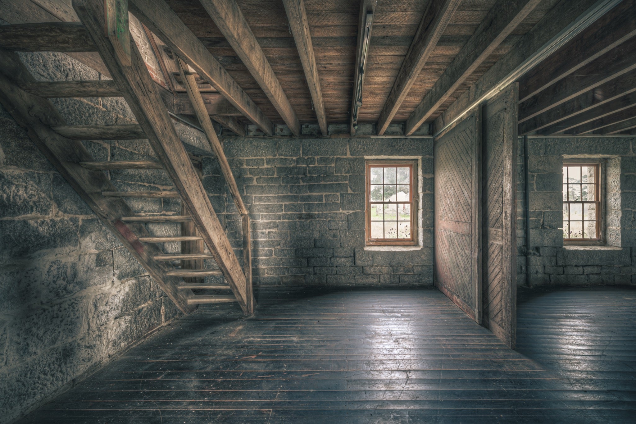 A wooden staircase rises along the stone wall of a timber-floored room at Jembaicumbene, with two double-hung windows in the rear wall looking out to open ground.
