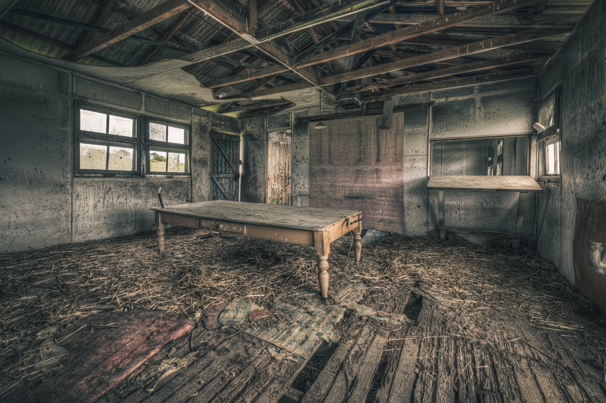 A long timber table with turned legs on a straw-strewn floor of broken boards, with multi-pane windows and an open plank door visible in a derelict interior at Jembaicumbene, New South Wales.