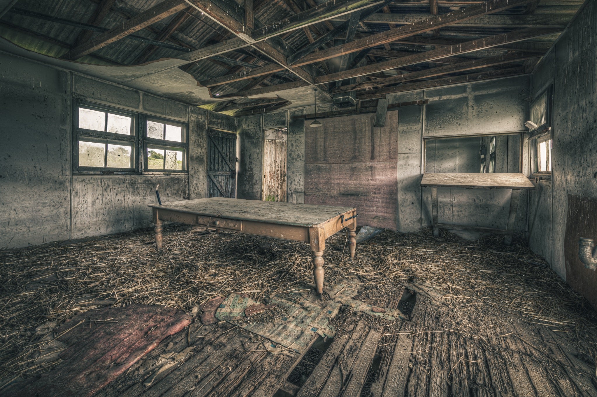A long timber table with turned legs on a straw-strewn floor of broken boards, with multi-pane windows and an open plank door visible in a derelict interior at Jembaicumbene, New South Wales.