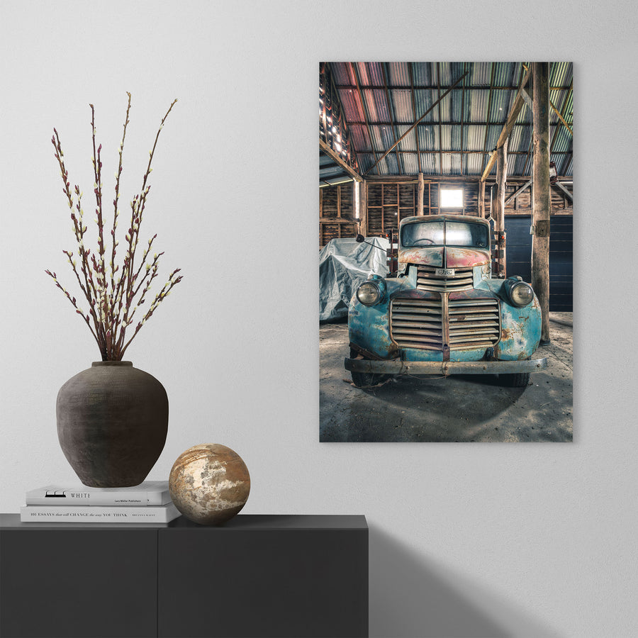 An old truck with a faded blue and rust body parked front-on inside the timber wagon barn at Jembaicumbene, its chrome grille and round headlights lit by a small window above.