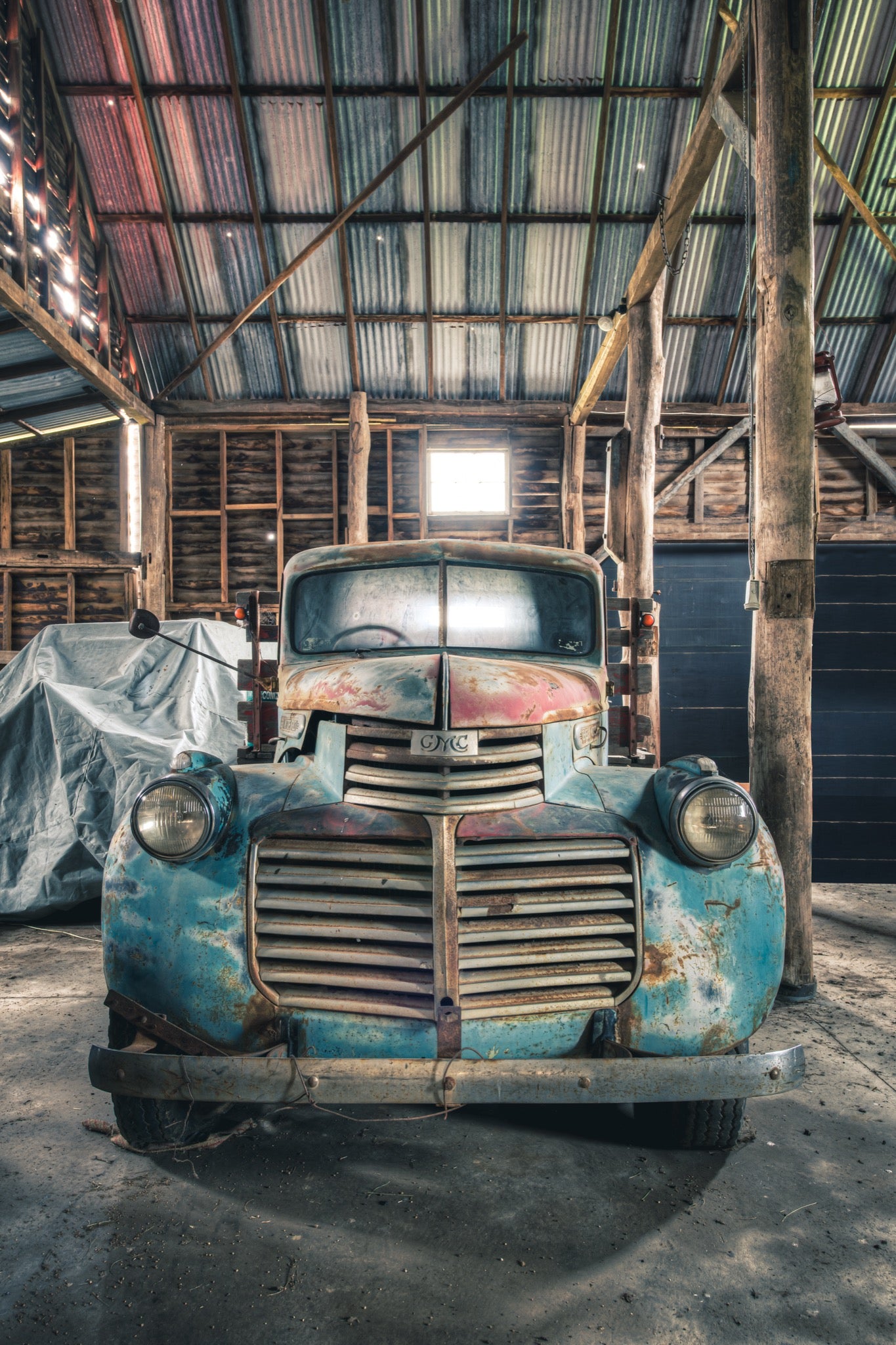 An old truck with a faded blue and rust body parked front-on inside the timber wagon barn at Jembaicumbene, its chrome grille and round headlights lit by a small window above.