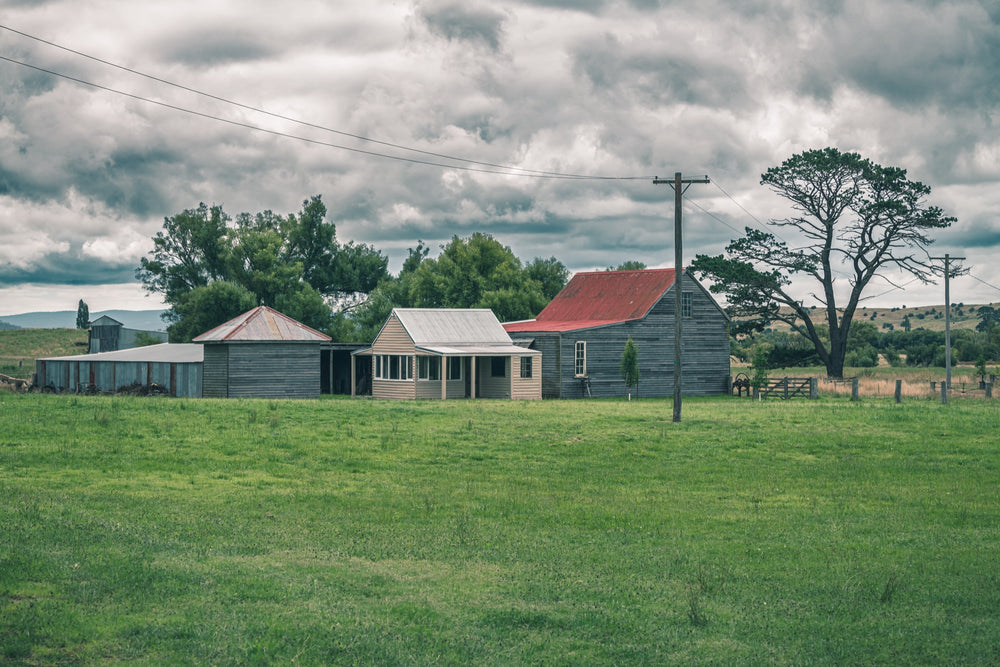 Farm buildings at Jembaicumbene, a red iron roofed barn and timber cottage across a green paddock.