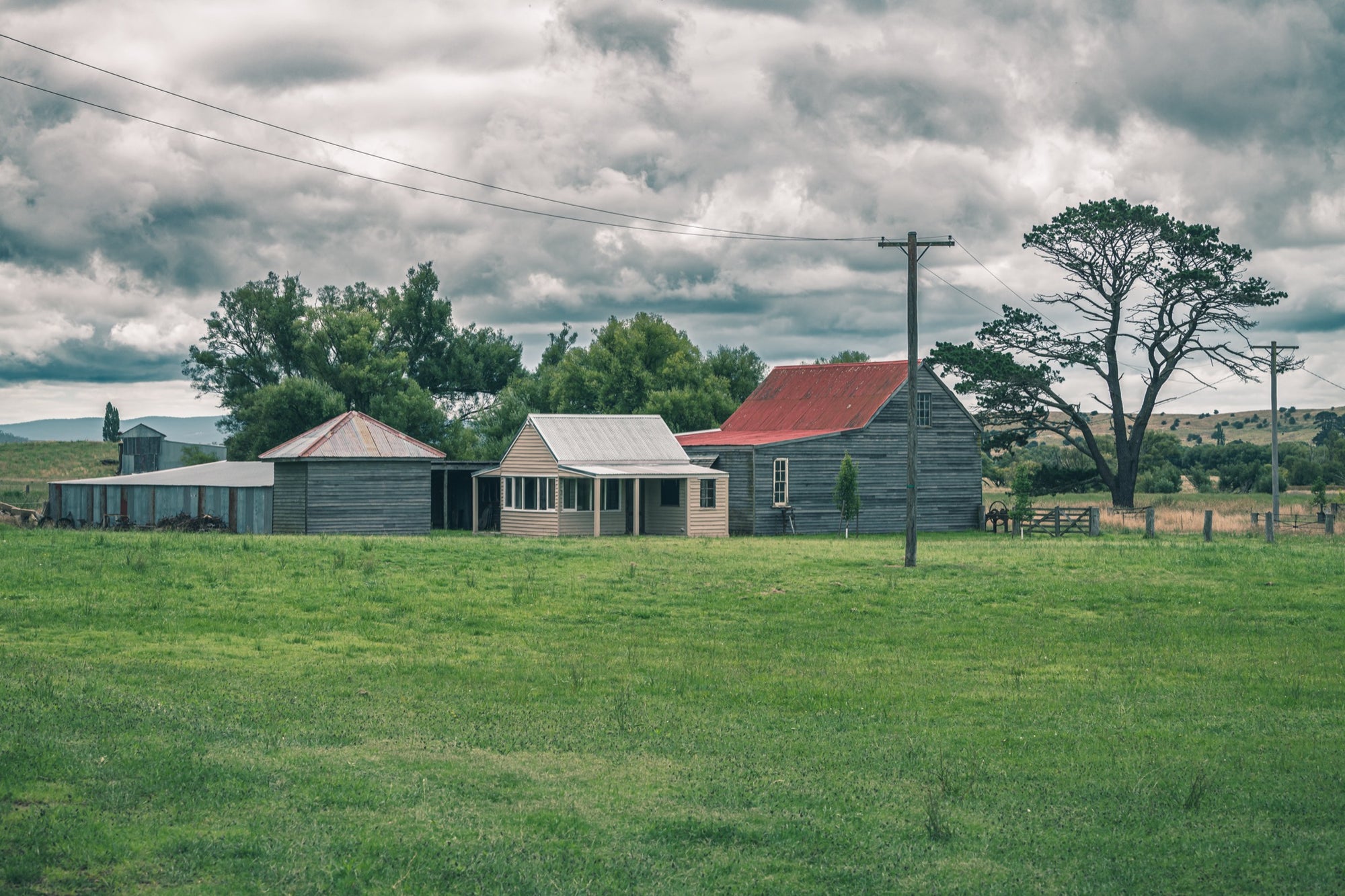 Farm buildings at Jembaicumbene, a red iron roofed barn and timber cottage across a green paddock.