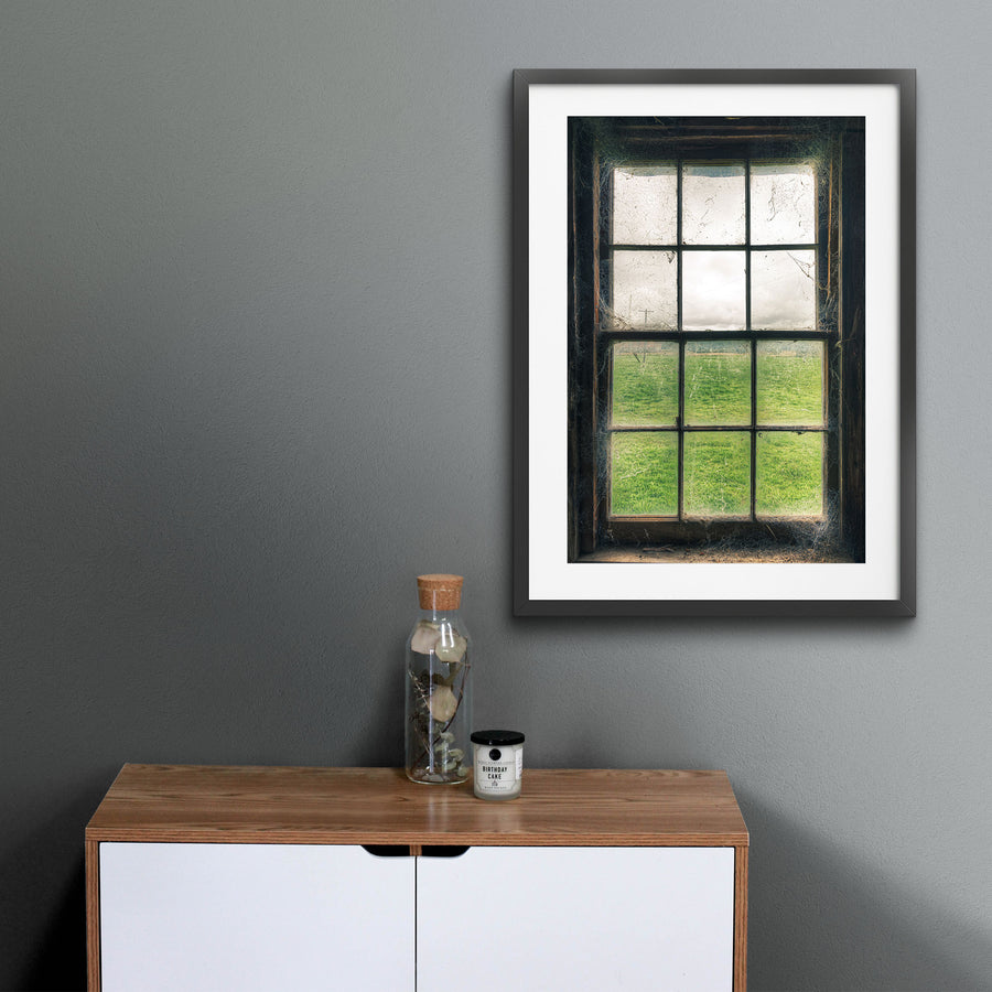 Timber sash window at Jembaicumbene, dusty panes and cobwebs framing a green paddock and overcast sky.