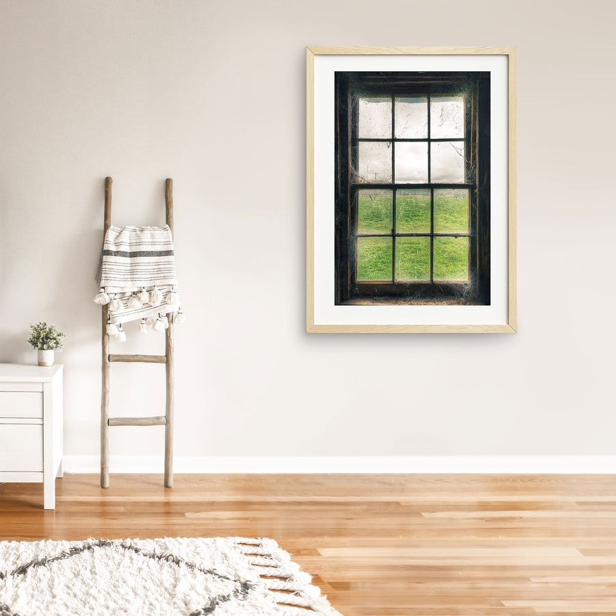 Timber sash window at Jembaicumbene, dusty panes and cobwebs framing a green paddock and overcast sky.