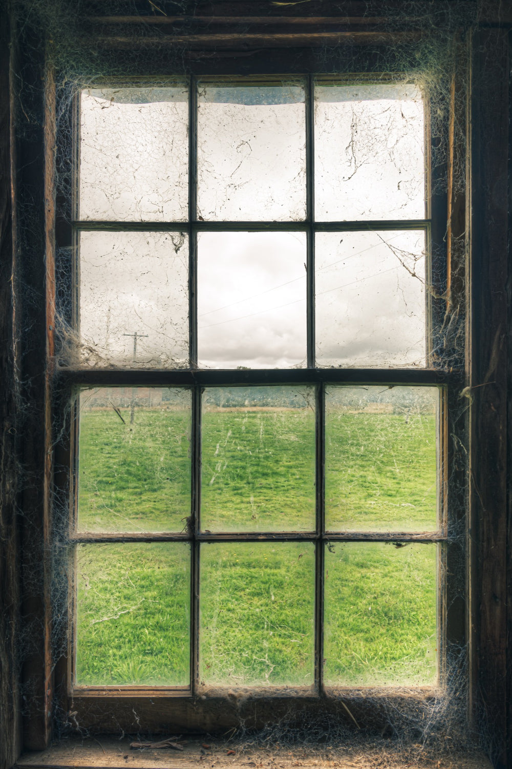Timber sash window at Jembaicumbene, dusty panes and cobwebs framing a green paddock and overcast sky.