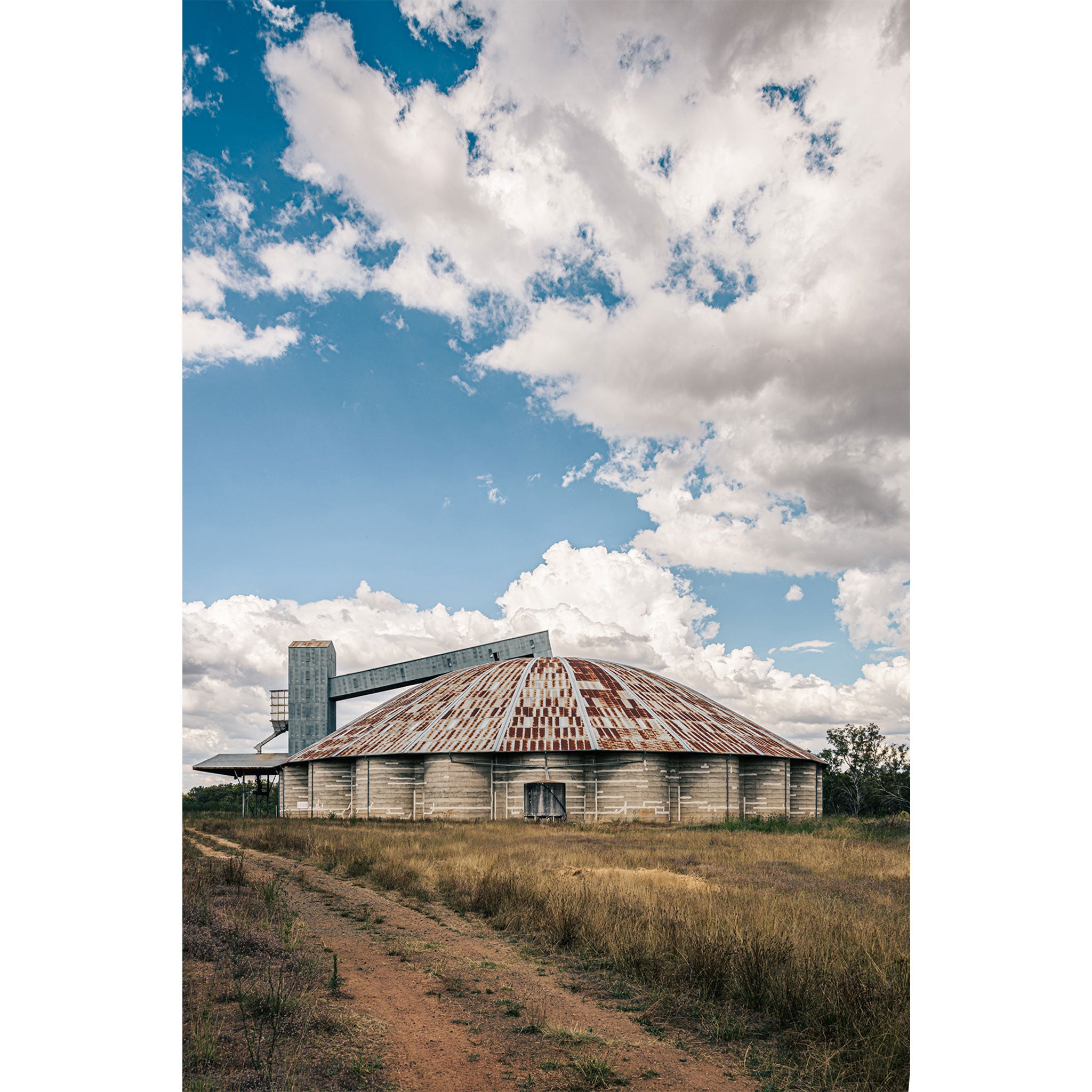 A rusted domed concrete grain silo with a raised conveyor gantry and headhouse tower at Mount Russell, set against a broad cloud-filled sky.