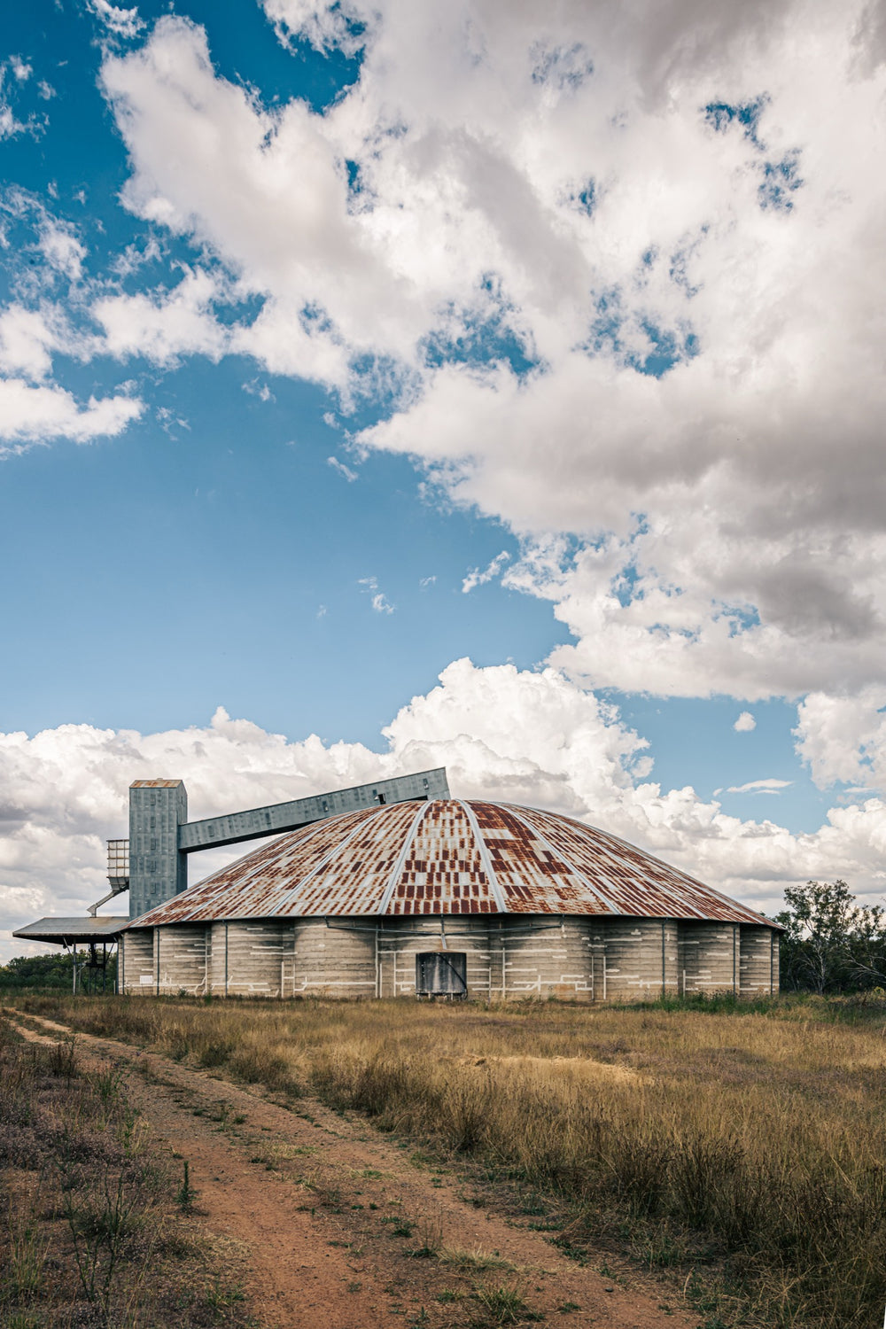 A rusted domed concrete grain silo with a raised conveyor gantry and headhouse tower at Mount Russell, set against a broad cloud-filled sky.