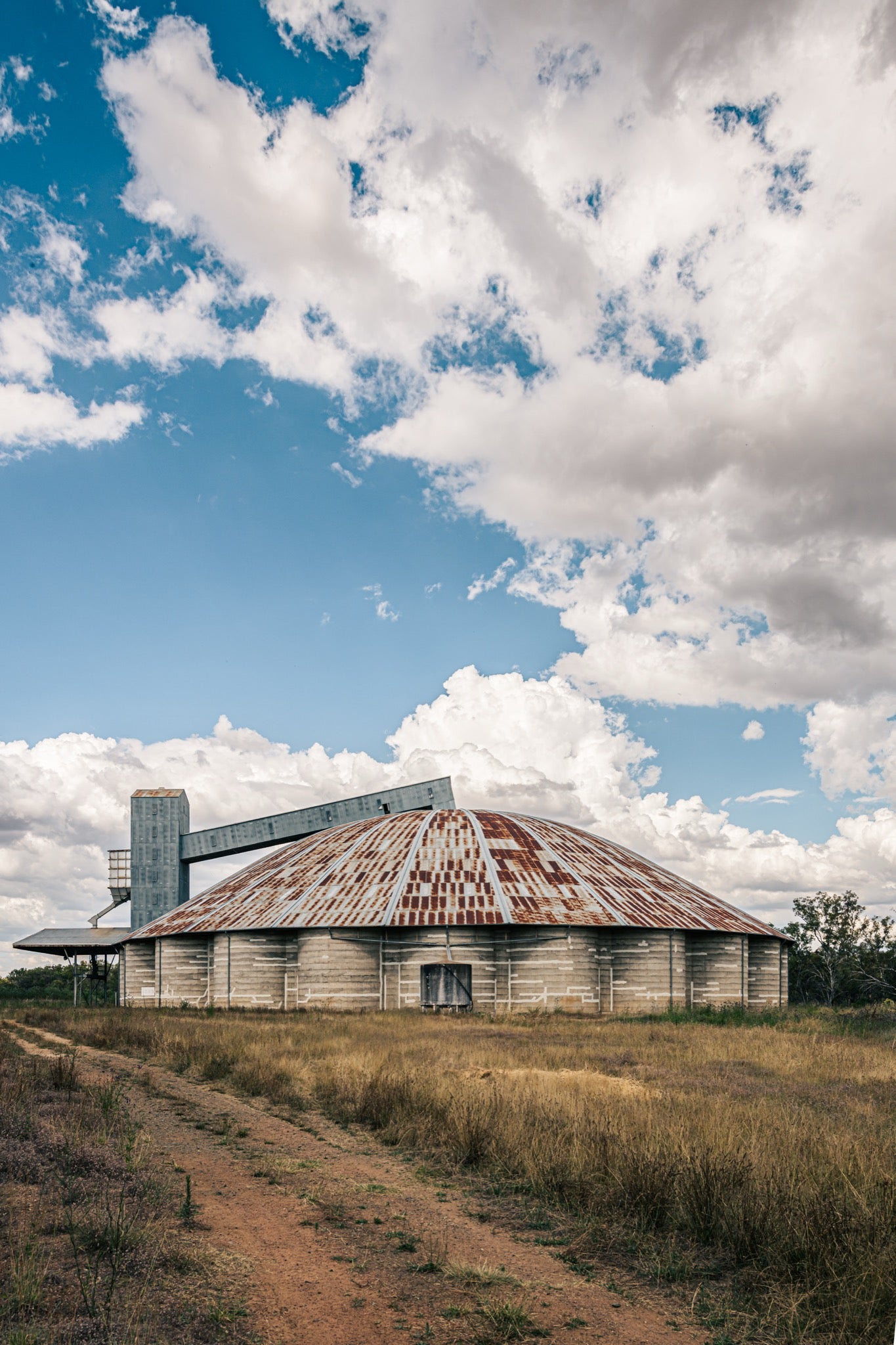A rusted domed concrete grain silo with a raised conveyor gantry and headhouse tower at Mount Russell, set against a broad cloud-filled sky.
