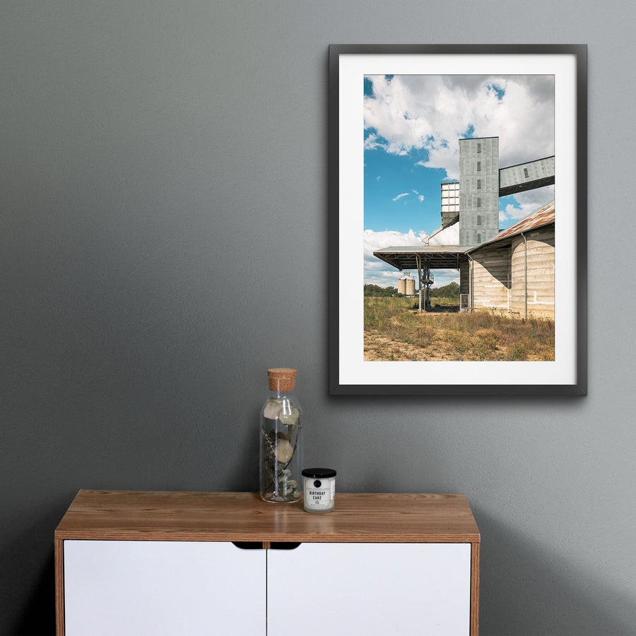 A tall corrugated metal grain elevator tower connected by a raised conveyor gallery and steel stairway to a concrete grain shed at Mount Russell, with smaller silos visible across a grassed paddock under a partly clouded sky.