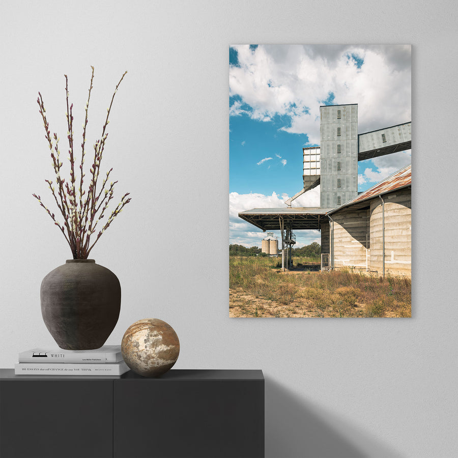 A tall corrugated metal grain elevator tower connected by a raised conveyor gallery and steel stairway to a concrete grain shed at Mount Russell, with smaller silos visible across a grassed paddock under a partly clouded sky.