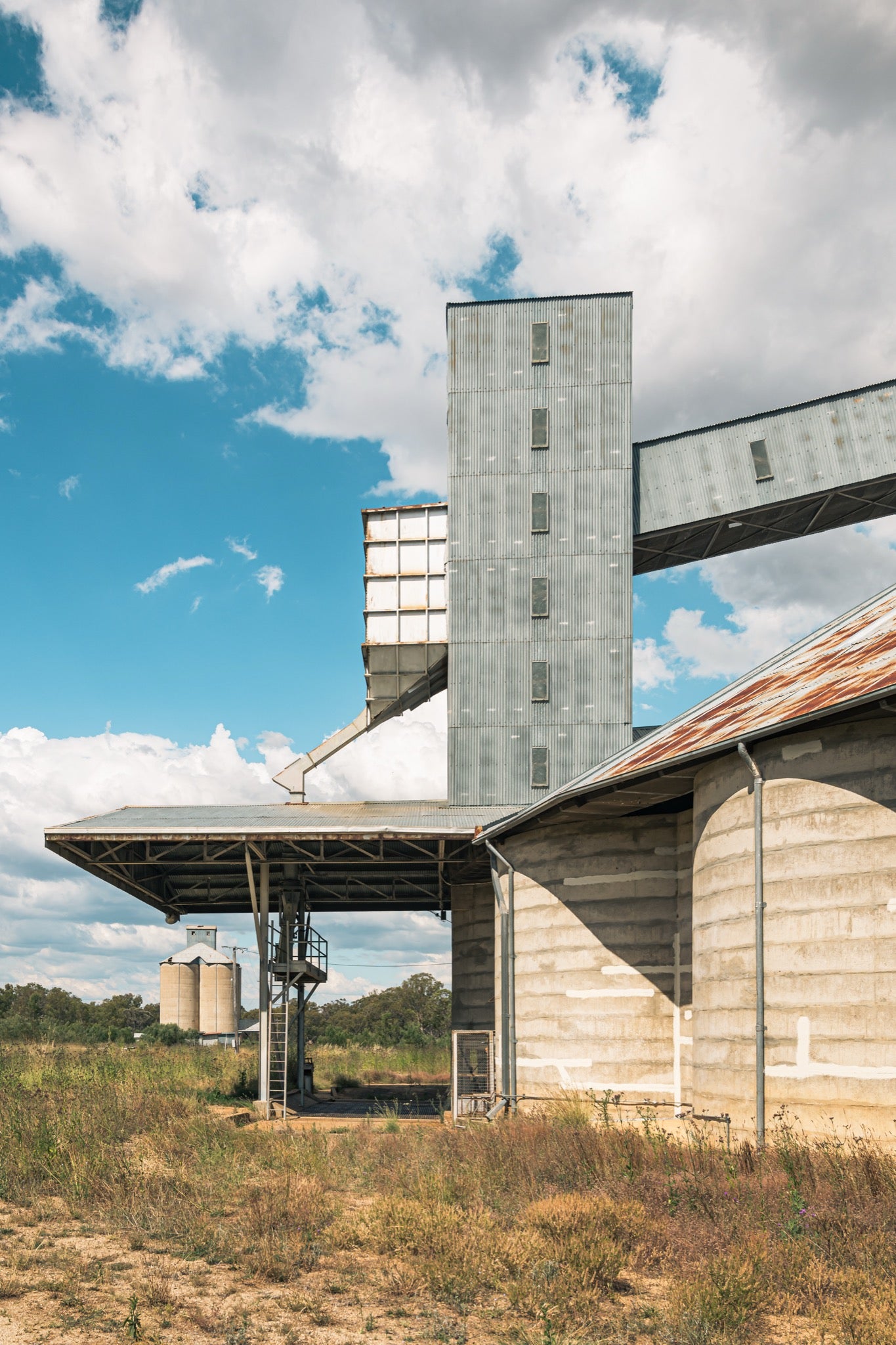 A tall corrugated metal grain elevator tower connected by a raised conveyor gallery and steel stairway to a concrete grain shed at Mount Russell, with smaller silos visible across a grassed paddock under a partly clouded sky.