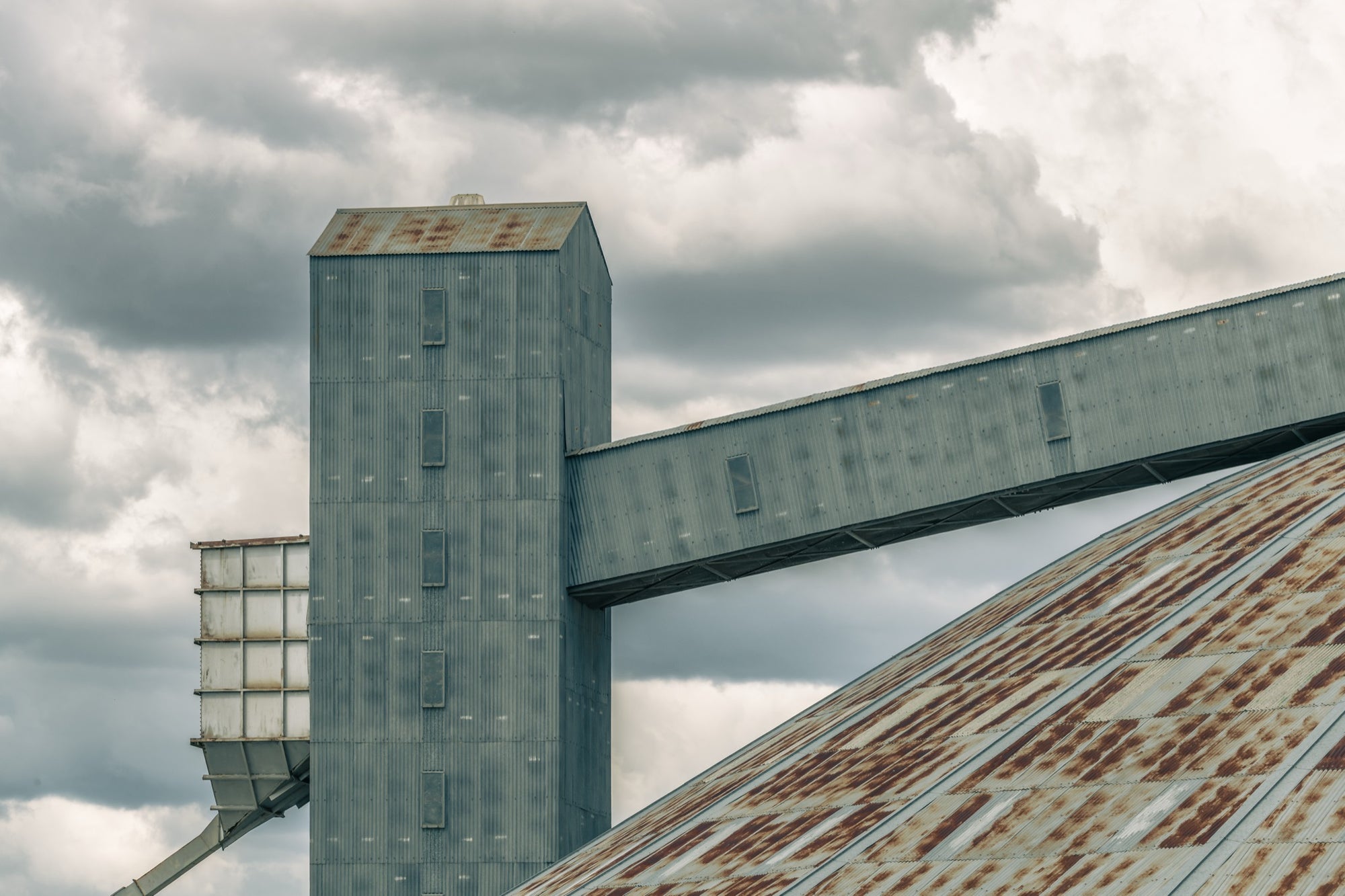A metal-clad tower and raised covered conveyor running to a rust-streaked pitched roof at Mount Russell, photographed under an overcast sky.