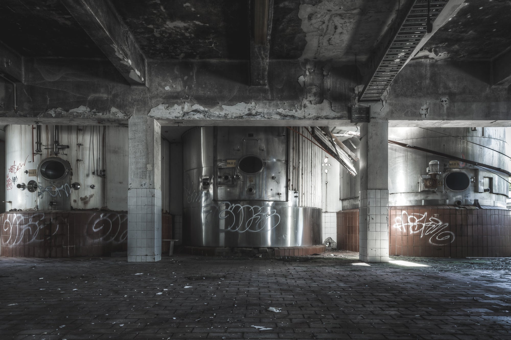 Raw Milk Silos at Peters Ice Cream Factory, stainless steel silos sit in bays divided by concrete columns, their circular hatches dark and open.
