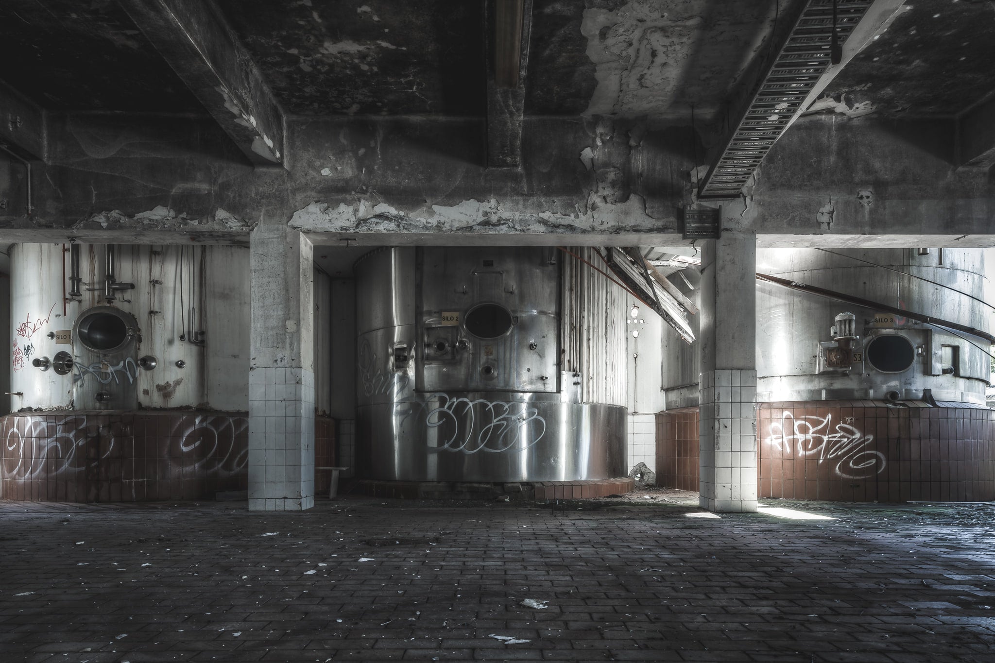 Raw Milk Silos at Peters Ice Cream Factory, stainless steel silos sit in bays divided by concrete columns, their circular hatches dark and open.