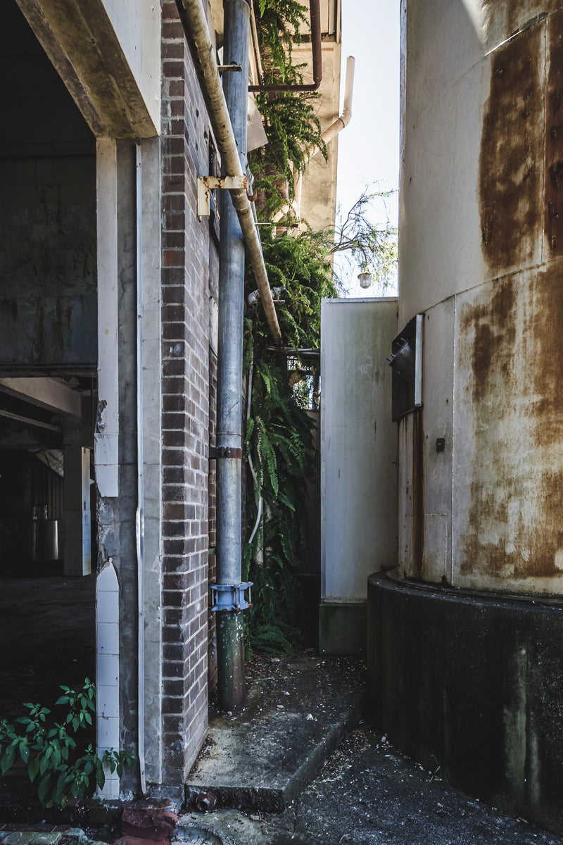 Water Vessel at Peters Ice Cream Factory, a narrow passage squeezes between a brick column and a curved concrete vessel stained deep ochre with rust.
