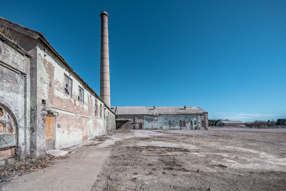 Workshop To Powerhouse at Portland Cement Works, a concrete path cuts across bare ground between two industrial buildings.