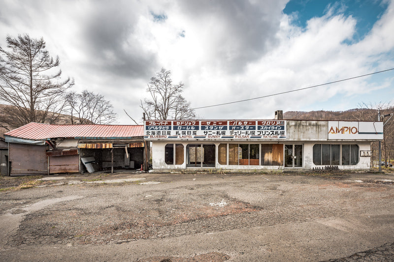 Faded model names, Bluebird, Cedric, Sunny, Pulsar and Gloria, run along a horizontal sign above broken windows on the former Nissan dealership in Yubari.