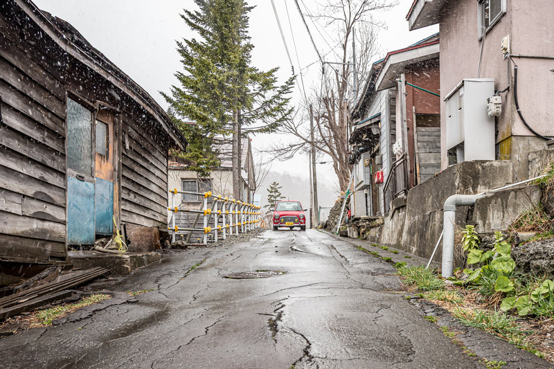 Snowflakes fall against dark utility poles and weathered timber-clad buildings along a sloping street in Yubari, the wet asphalt cracked and glistening under a grey sky.