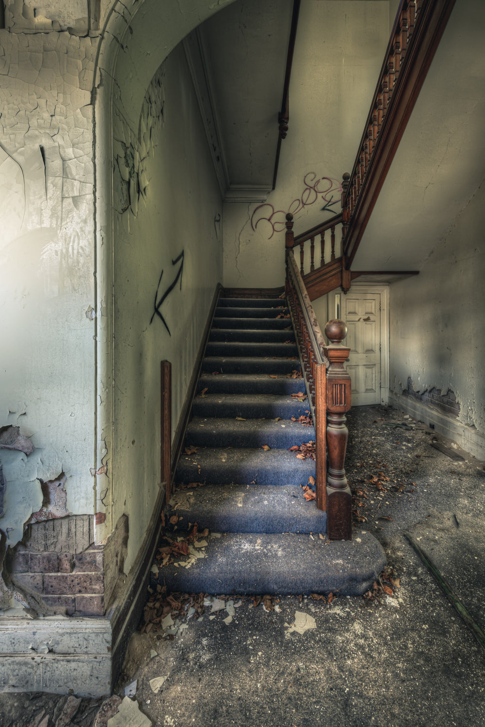 Crumbling Stairwell at The Asylum, a carved timber newel post stands at the base of a carpeted staircase.