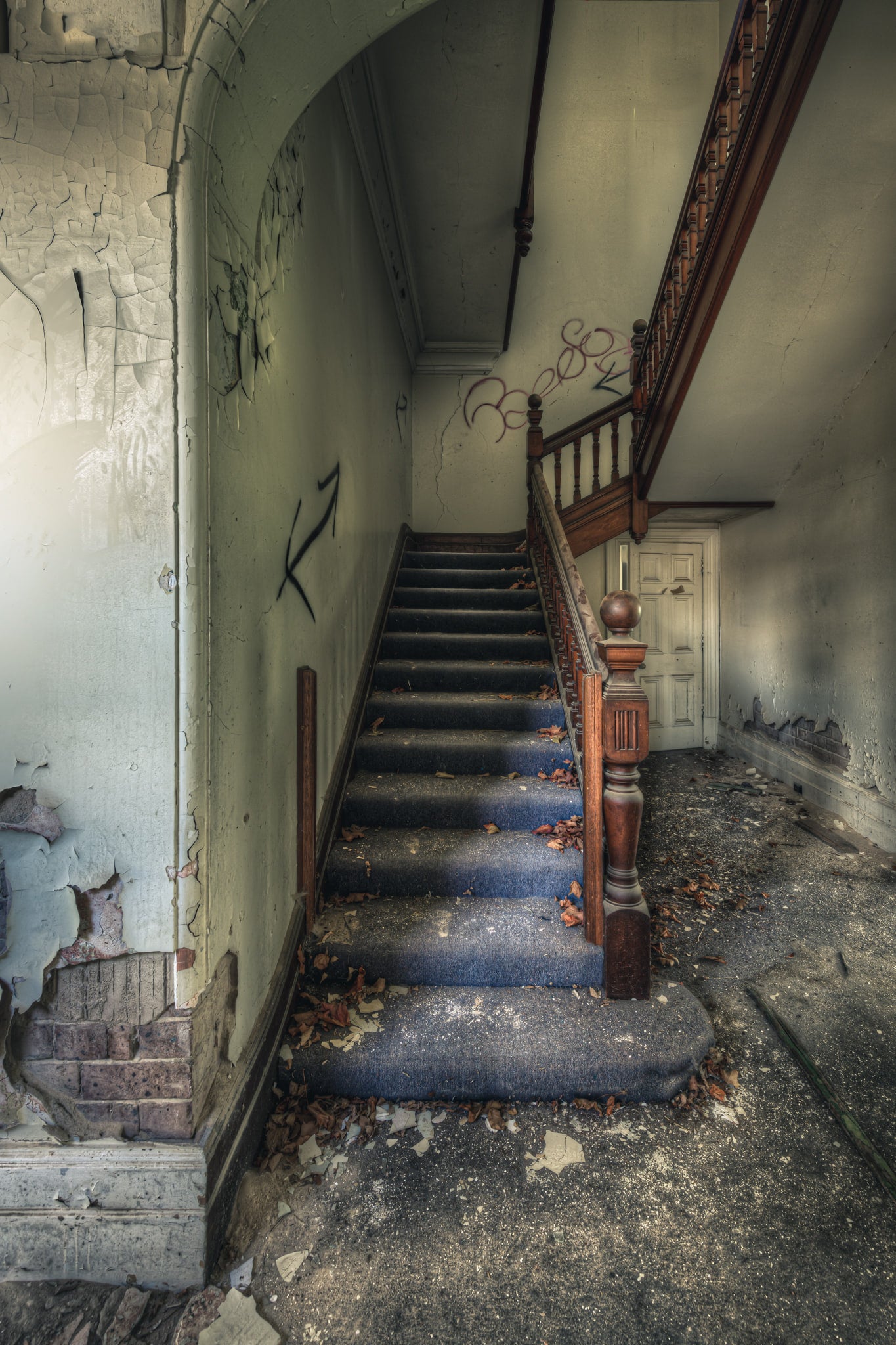 Crumbling Stairwell at The Asylum, a carved timber newel post stands at the base of a carpeted staircase.